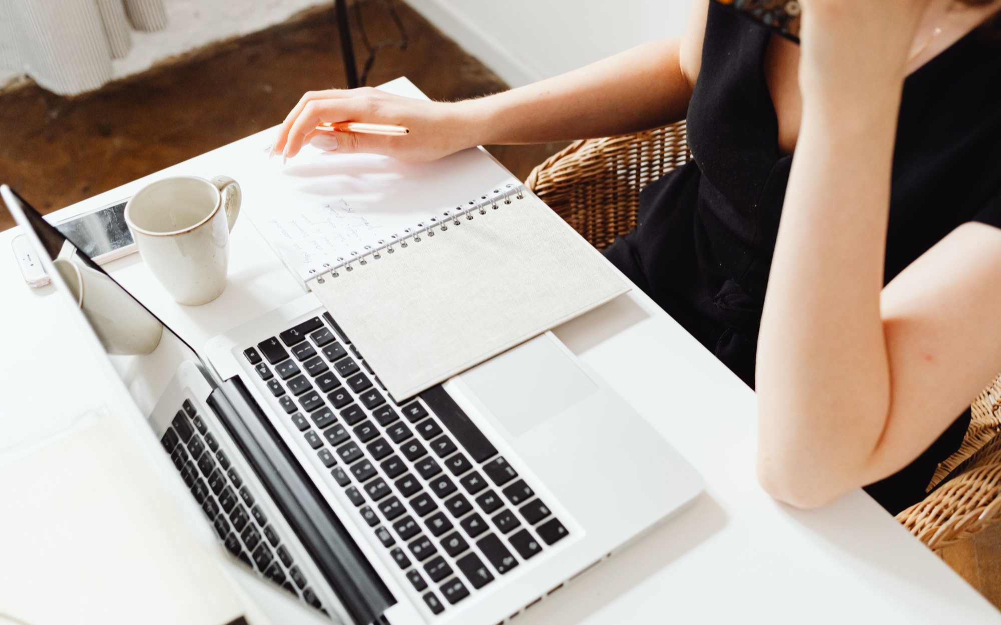 Frau sitzt an einem weißen Schreibtisch mit Laptop, Notizbuch und Kaffeetasse und schreibt Notizen