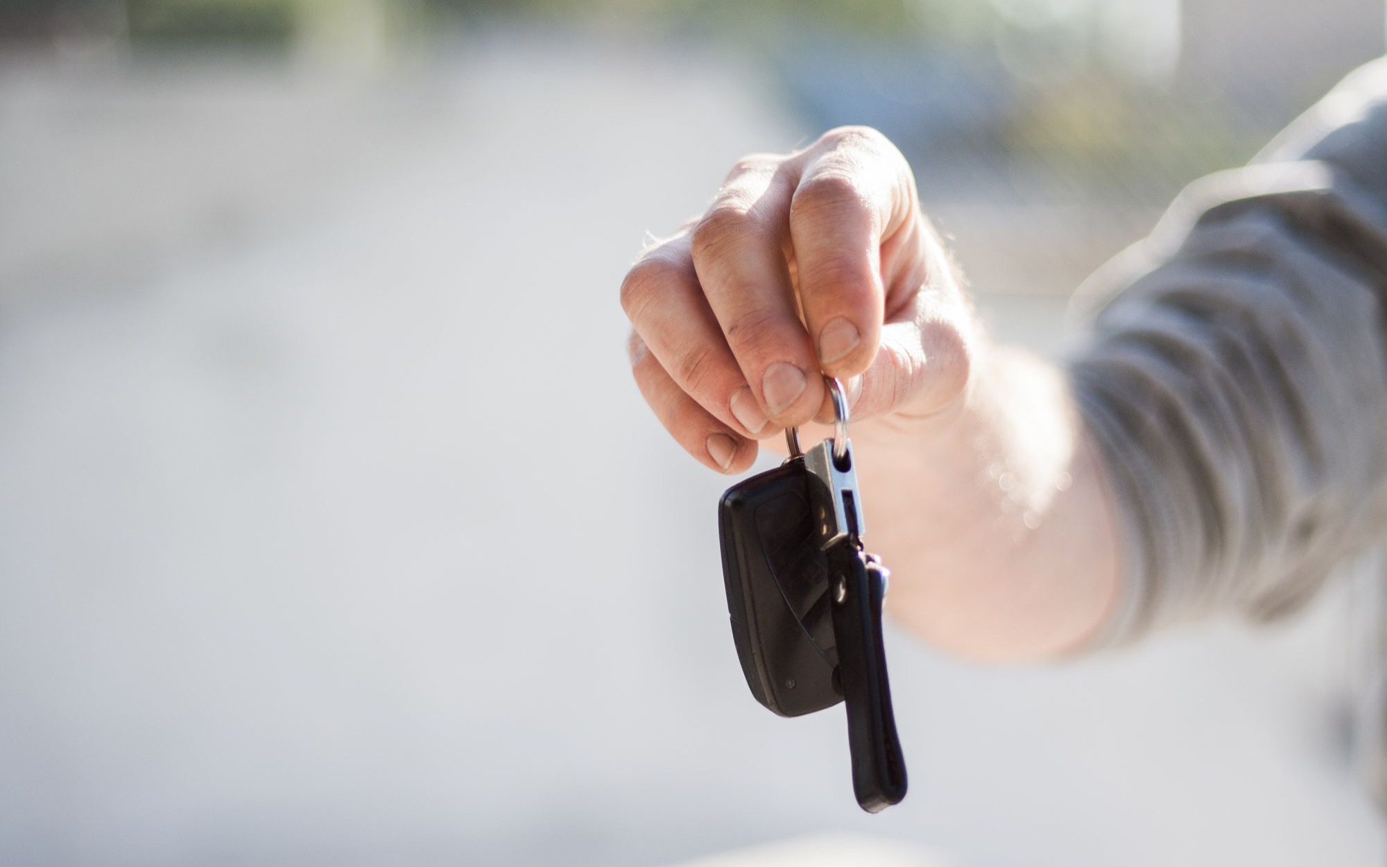 Hand reicht einen Autoschlüssel mit schwarzem Anhänger über einen hellen, unscharfen Hintergrund