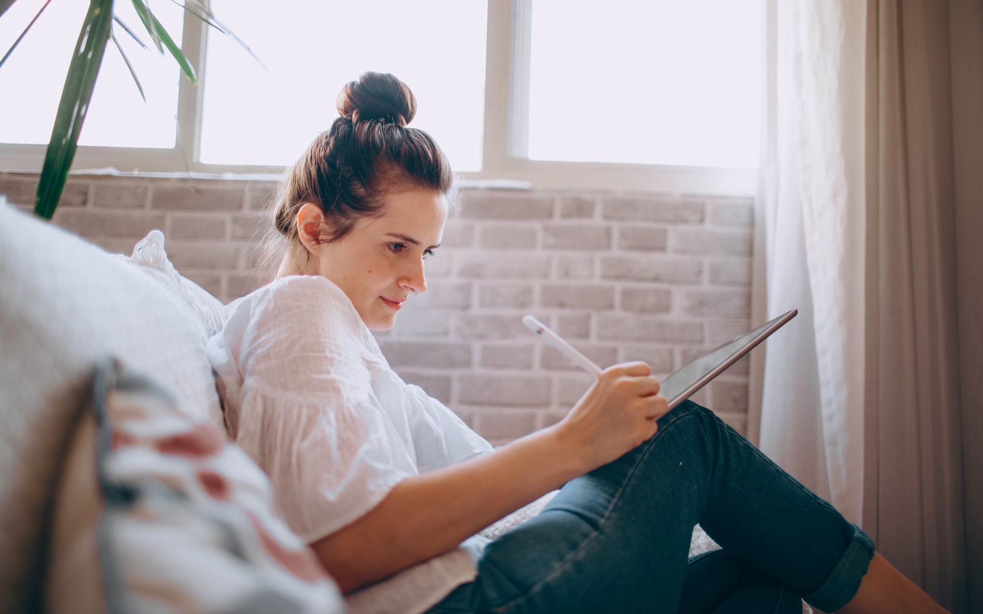 Junge Frau sitzt am Fenster auf einem Sofa und schreibt mit einem Stift auf einem Tablet