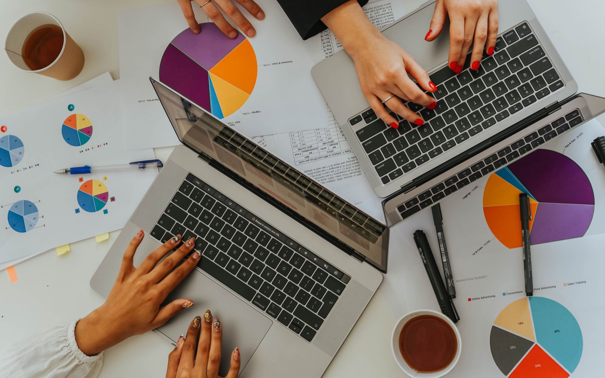 Mehrere Hände arbeiten an zwei Laptops zwischen ausgedruckten Kreisdiagrammen und Kaffeebechern