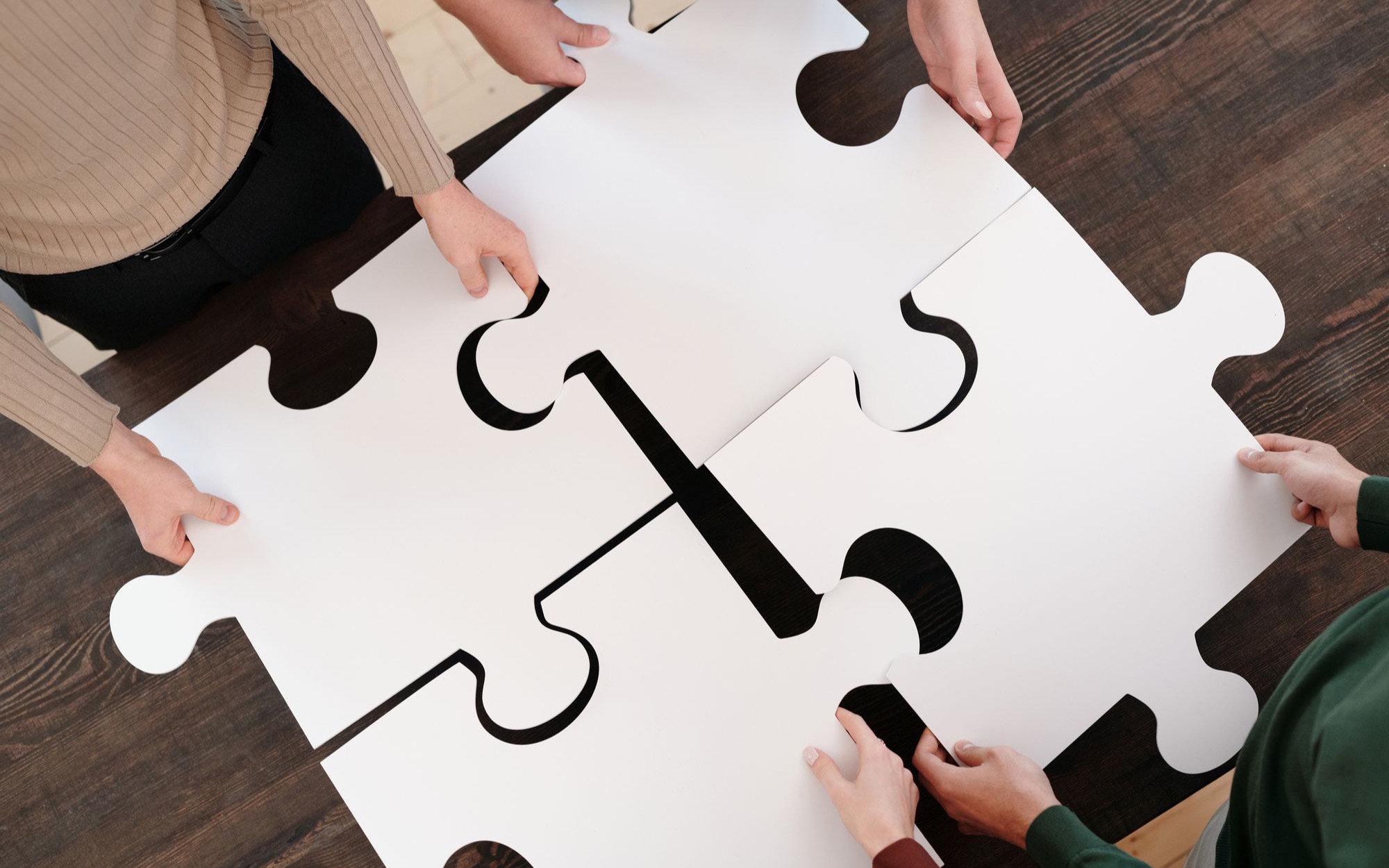 Mehrere Hände legen große weiße Puzzleteile auf einem Holztisch zusammen