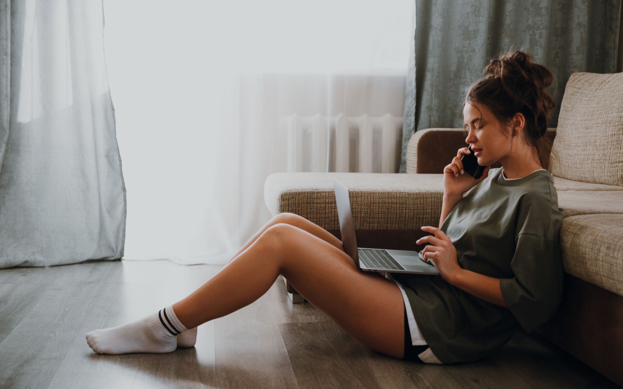 Junge Frau sitzt auf dem Boden vor einem Sofa, telefoniert und tippt auf einem Laptop