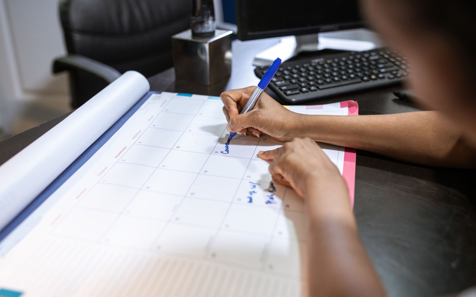 Hand trägt mit blauem Filzstift Termine in einen großen Monatskalender auf dem Schreibtisch ein