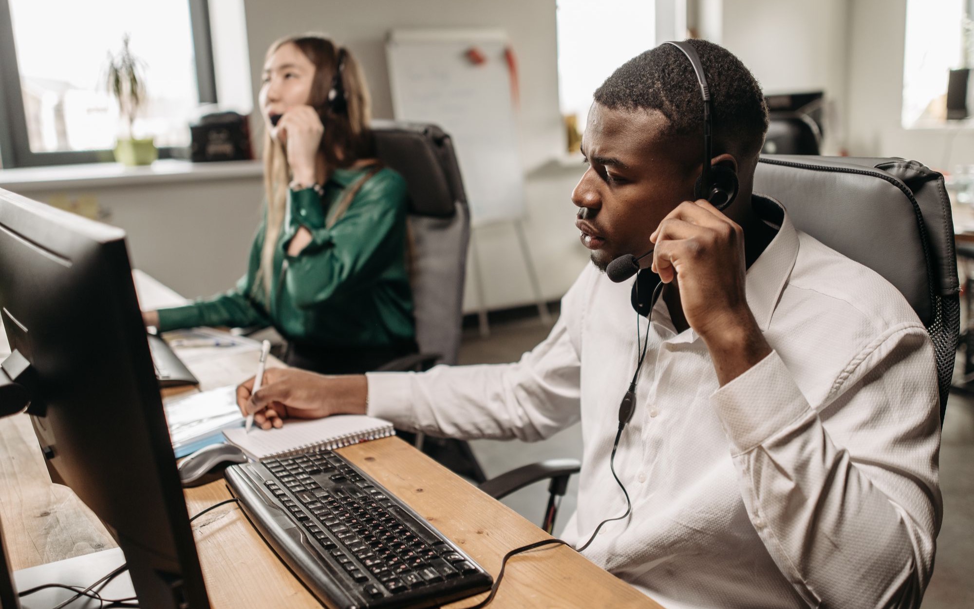 Mitarbeiter mit Headset telefoniert am Computer, im Hintergrund eine Kollegin am Telefon