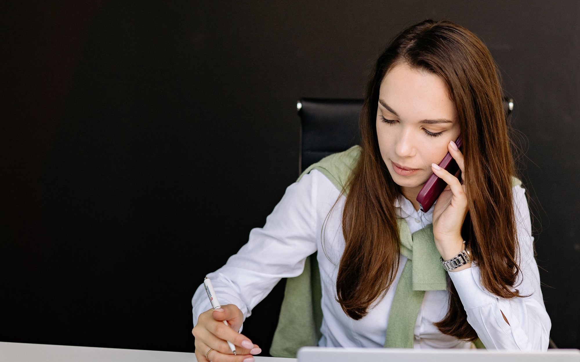 Ženska za pisalno mizo telefonira z mobilnikom in si ob prenosniku dela zapiske