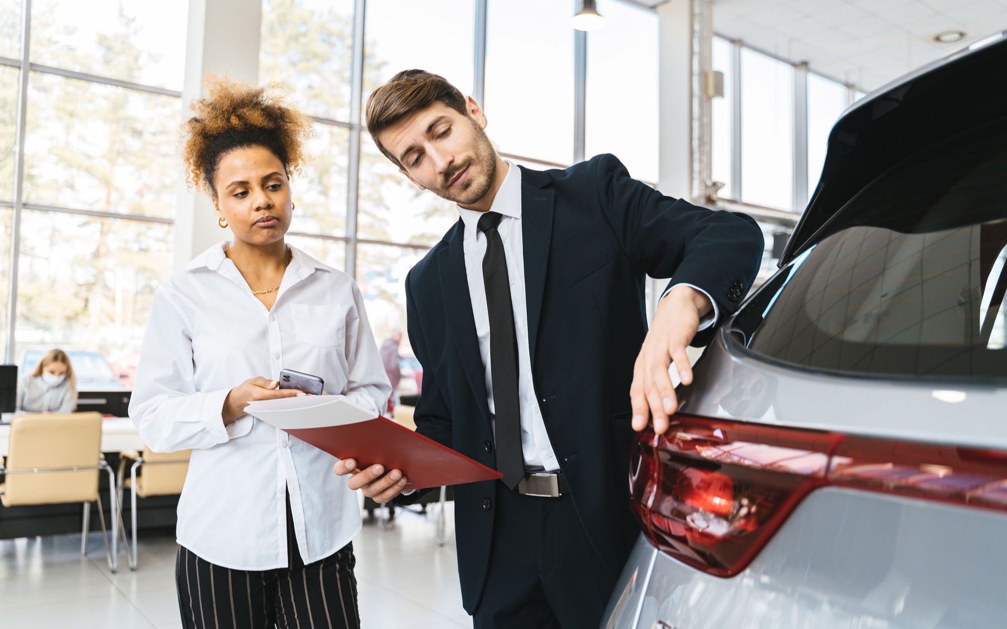 Verkäufer im Anzug zeigt einer Kundin mit Mappe ein Auto in einem hellen Autohaus
