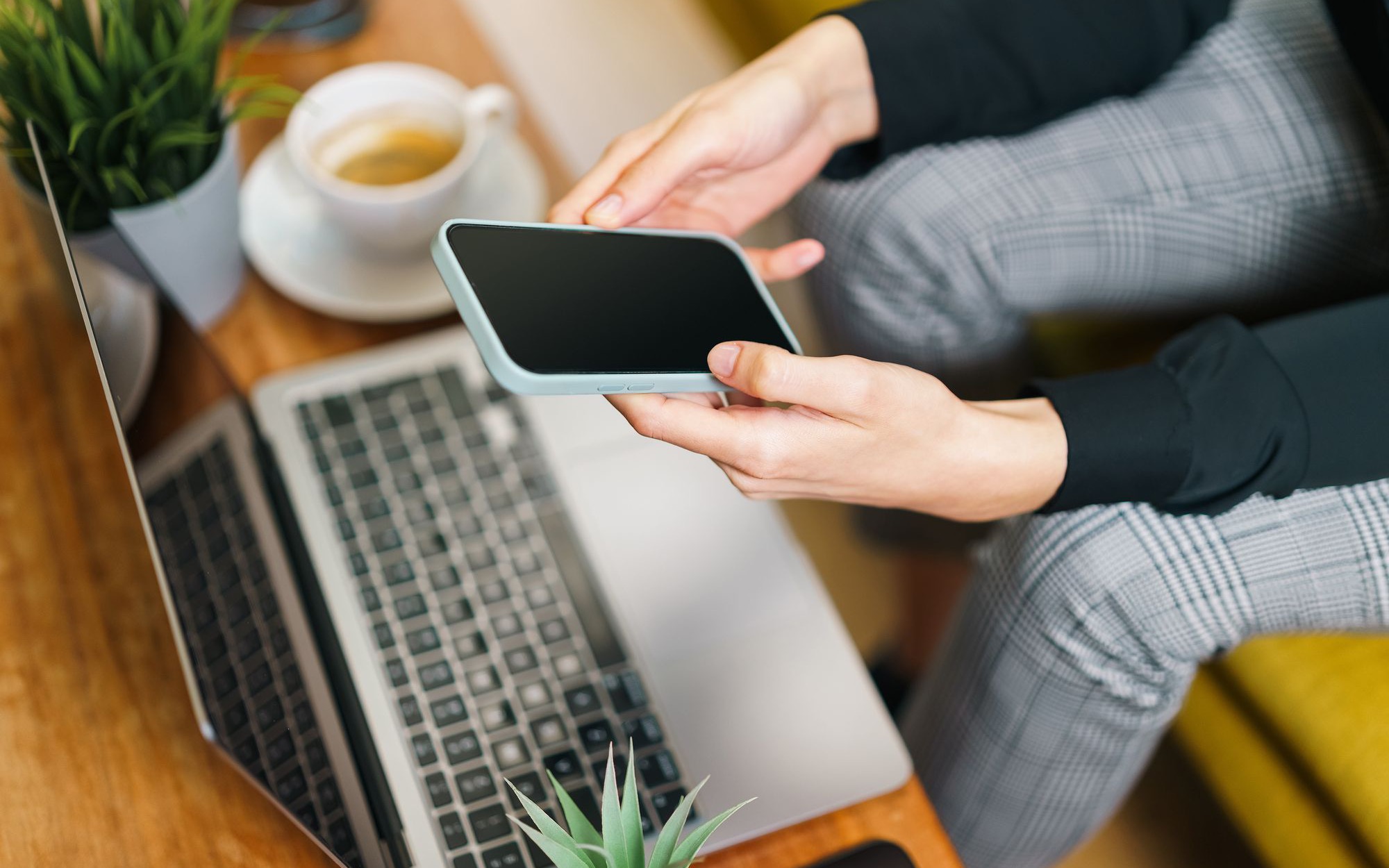 Hände halten ein Smartphone über einem Laptop, daneben eine Tasse Kaffee und Pflanzen
